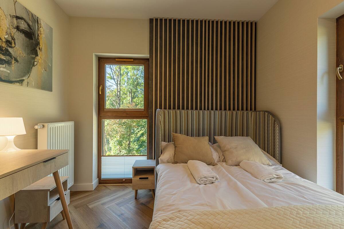 A bedroom with a bed, a desk, a radiator, and a window with a view of trees.