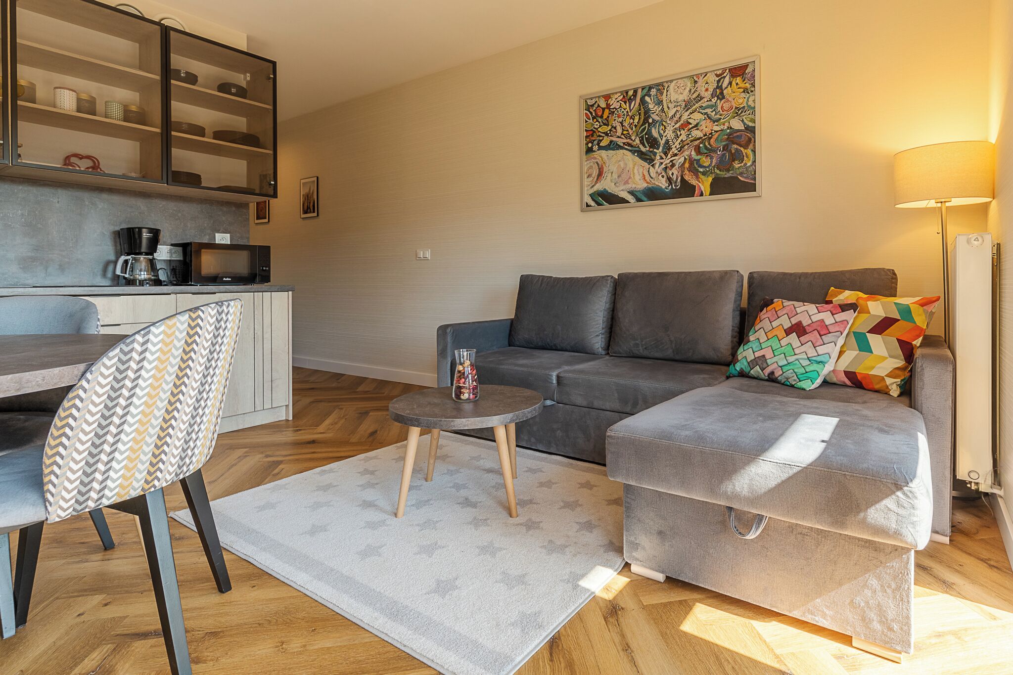 A modern living room with a grey sofa, a patterned armchair, a coffee table, and a kitchen area with a microwave and coffee maker.