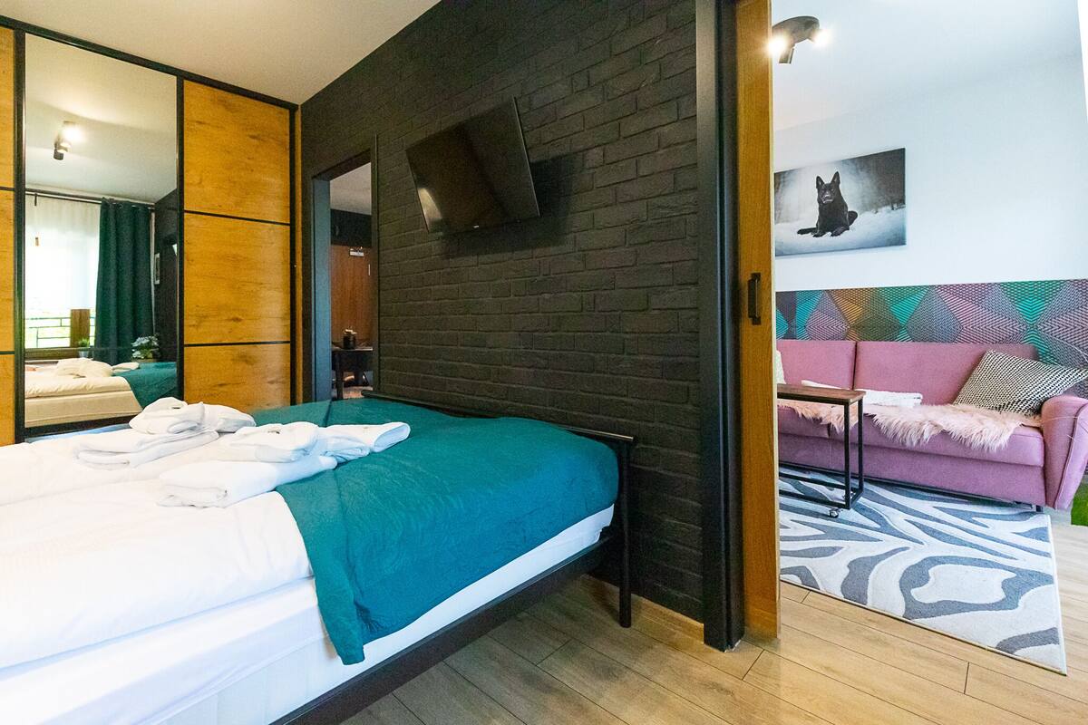 A modern bedroom with a bed, a large mirror, a TV mounted on the wall, and a pink sofa in the adjacent living area.