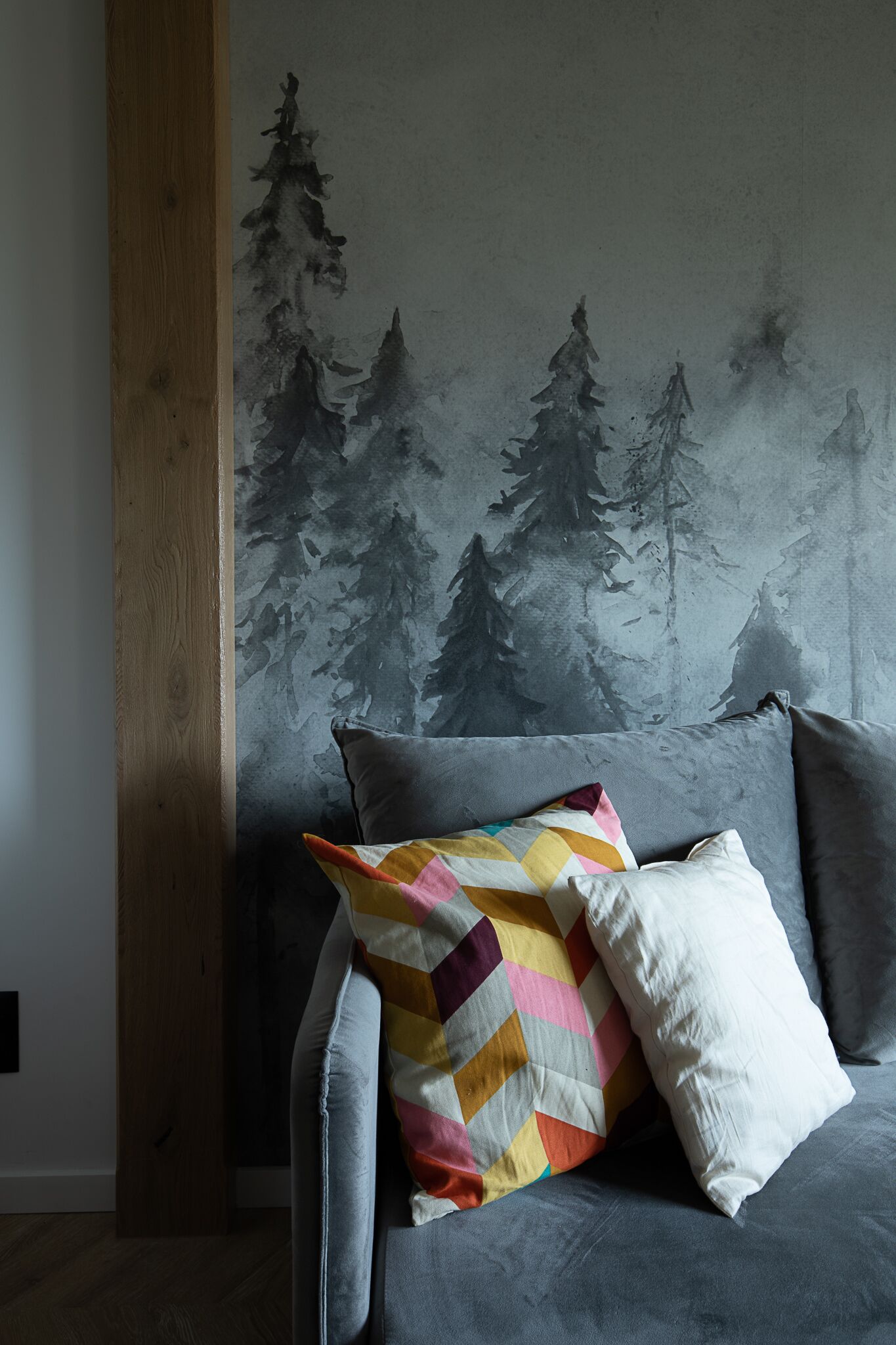 A grey sofa with a colorful quilted pillow and a white pillow, against a wall with a forest mural.