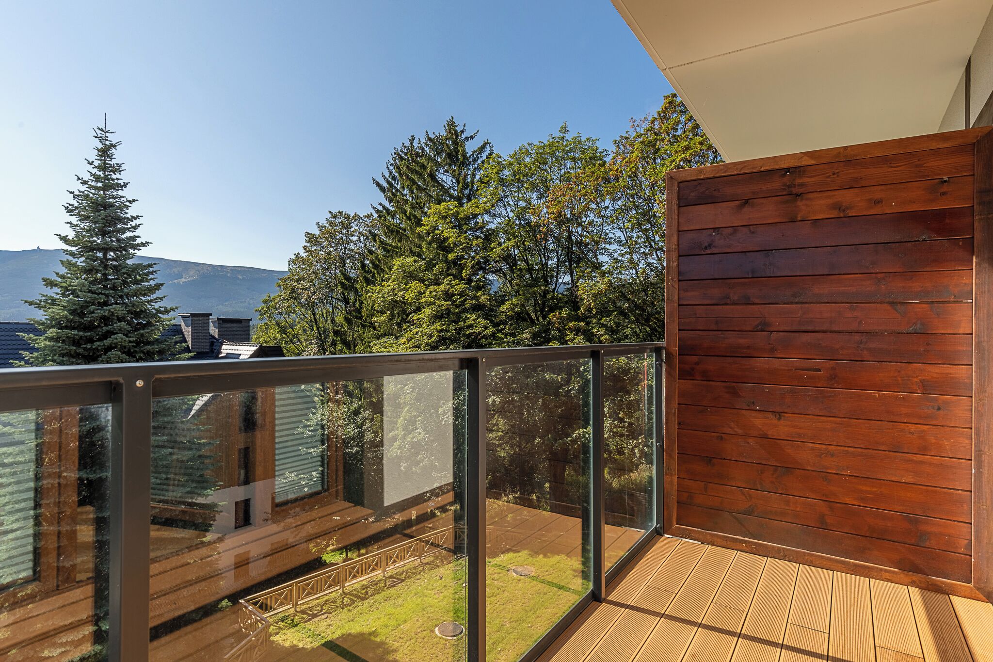 A balcony with a glass railing, wooden deck, and a view of trees and mountains.
