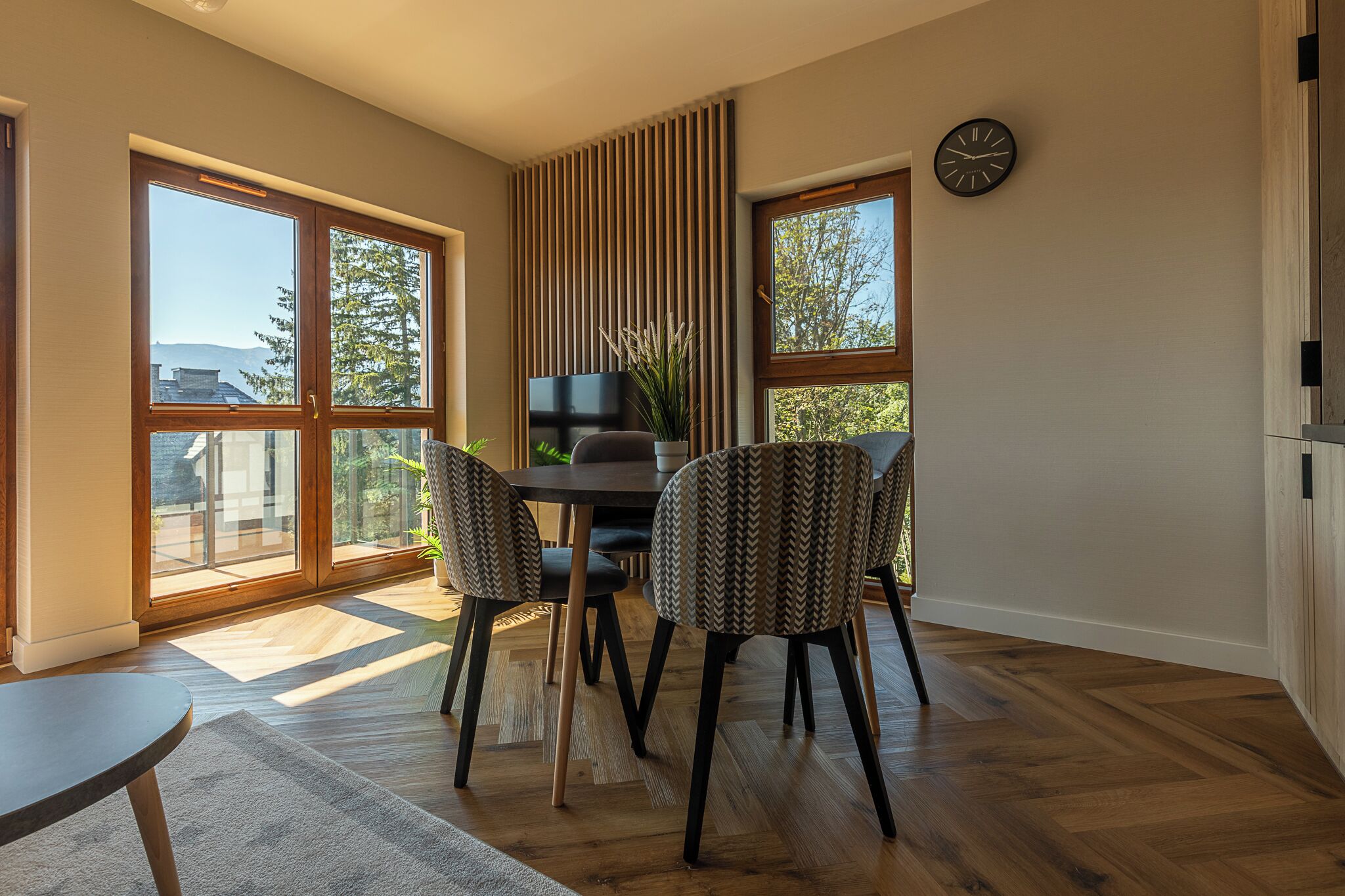 A dining area with a table and chairs, large windows, and a clock on the wall.