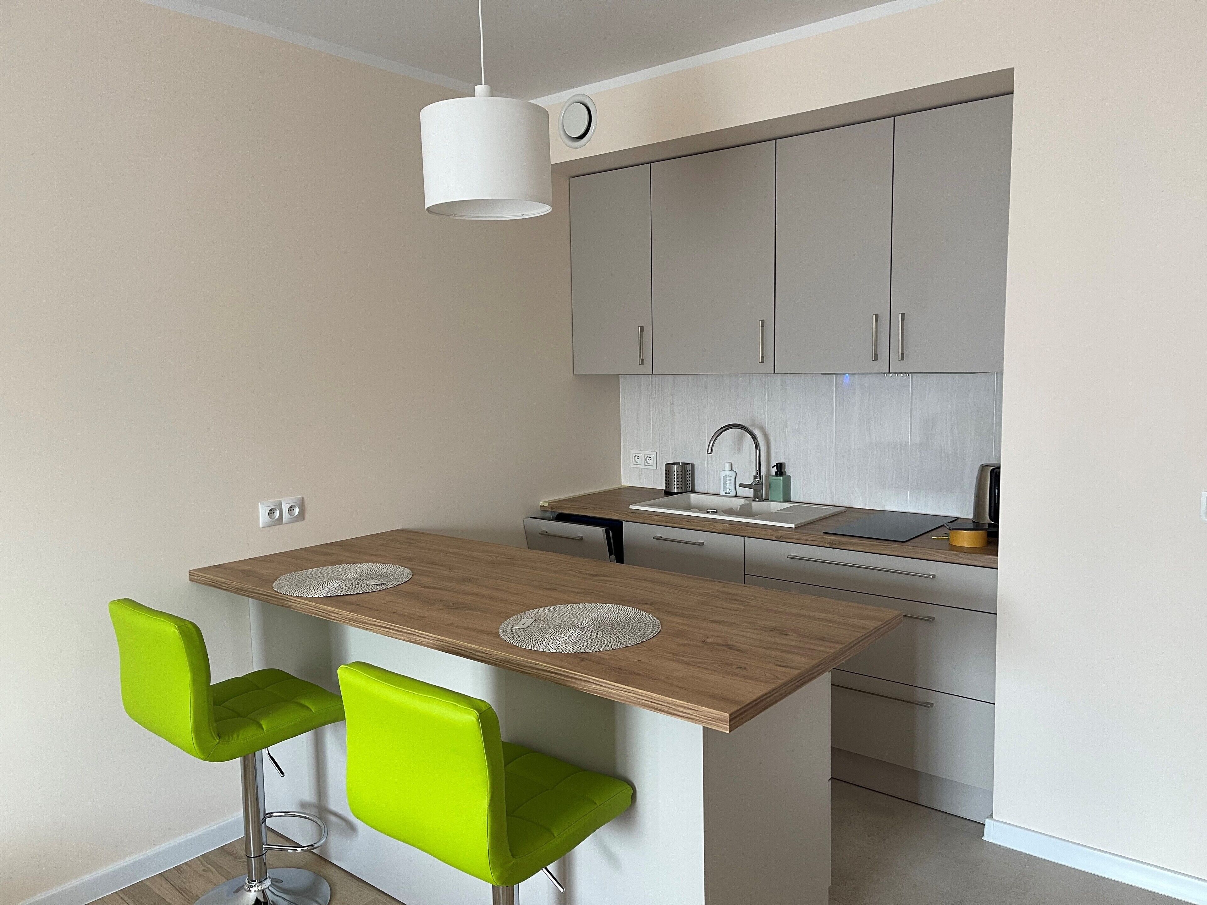 Una cocina compacta con una isla de madera, dos sillas verdes, una lámpara colgante blanca y muebles de cocina modernos.