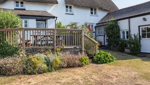 A traditional white house with a thatched roof, a wooden deck, and a garden with various plants and flowers. - End Cottage (Kingsbridge)