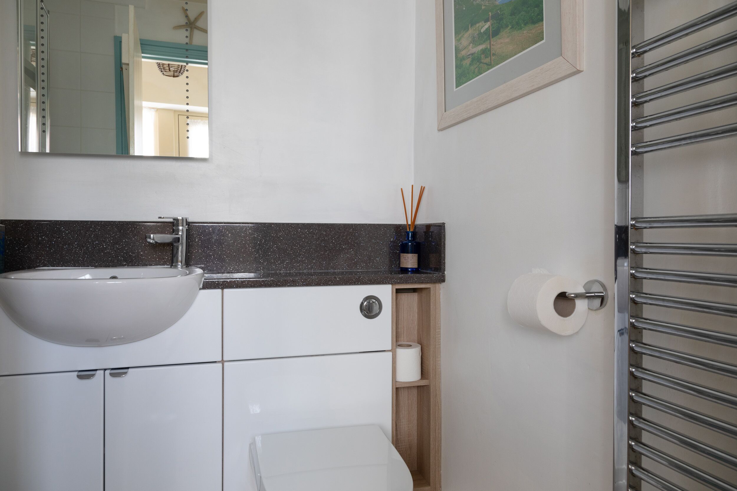 Un baño con lavamanos blanco, un espejo, un inodoro, un perchero para toallas y un cuadro en la pared.