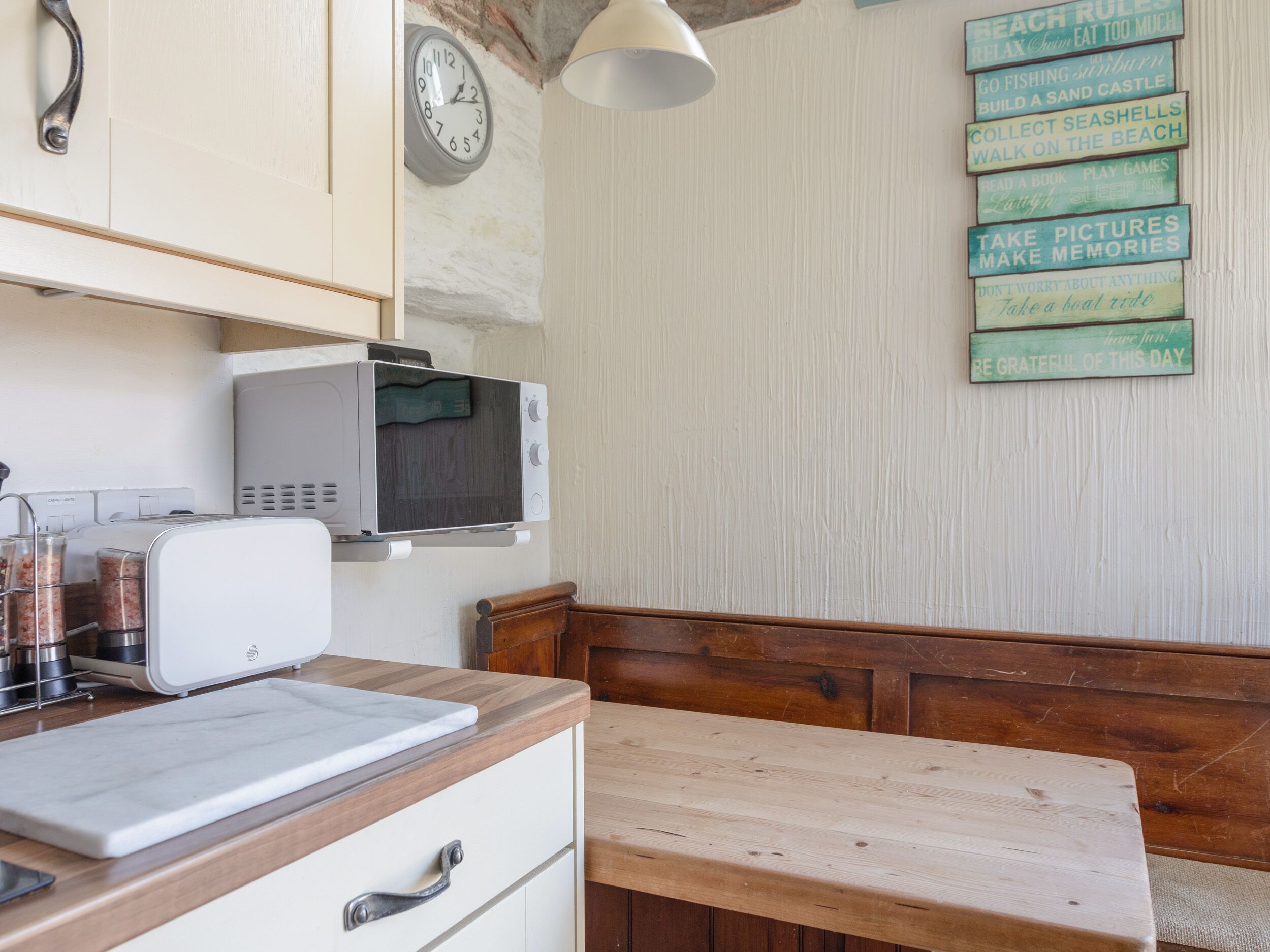 A kitchen with a microwave, a clock, and a wooden bench.