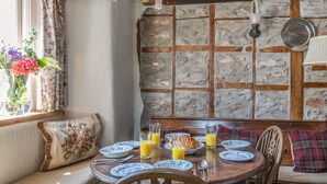 A cozy dining area with a stone wall, wooden beams, and a window with floral curtains. - End Cottage (Kingsbridge)