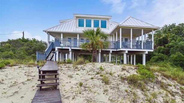 A beachfront house with a wooden deck leading to a balcony, surrounded by sand and vegetation.