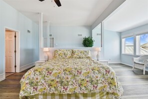 Une chambre à coucher avec une couvre-lit à motifs floraux, une table de chevet blanche, une plante en pot et une vue sur le ciel par la fenêtre.