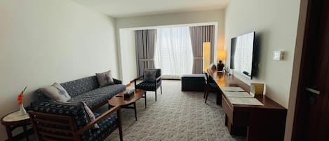 A hotel room with a sofa, armchair, ottoman, a small table, a desk, a TV, and a window with curtains.