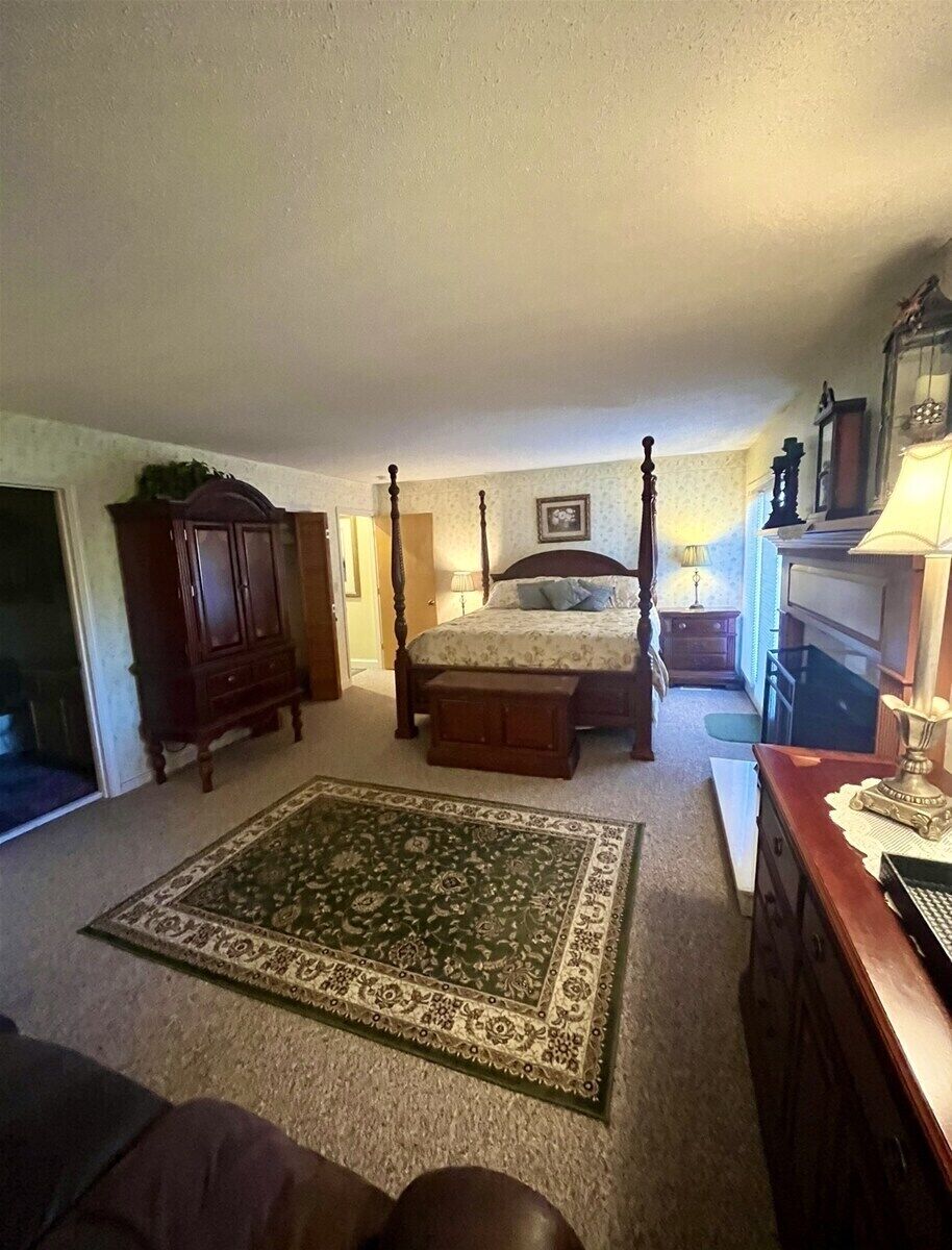 A bedroom with a four-poster bed, a wooden dresser, a nightstand, a lamp, and a large area rug.