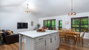 Cocina amplia con una isla central, mesa de comedor de madera y sillas. Un ventanal grande ofrece vistas a zonas verdes.