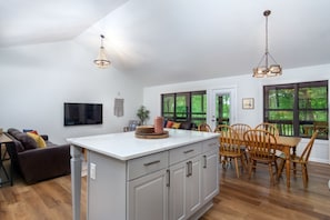 A spacious kitchen with a central island, wooden dining table, and chairs. There is a large window providing a view of greenery outside. - Turkey Run at Rumbling Bald (Lake Lure)