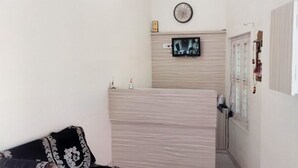Une petite pièce, de taille modeste, comprenant un lit, un téléviseur fixé au mur et une horloge placée au-dessus.
