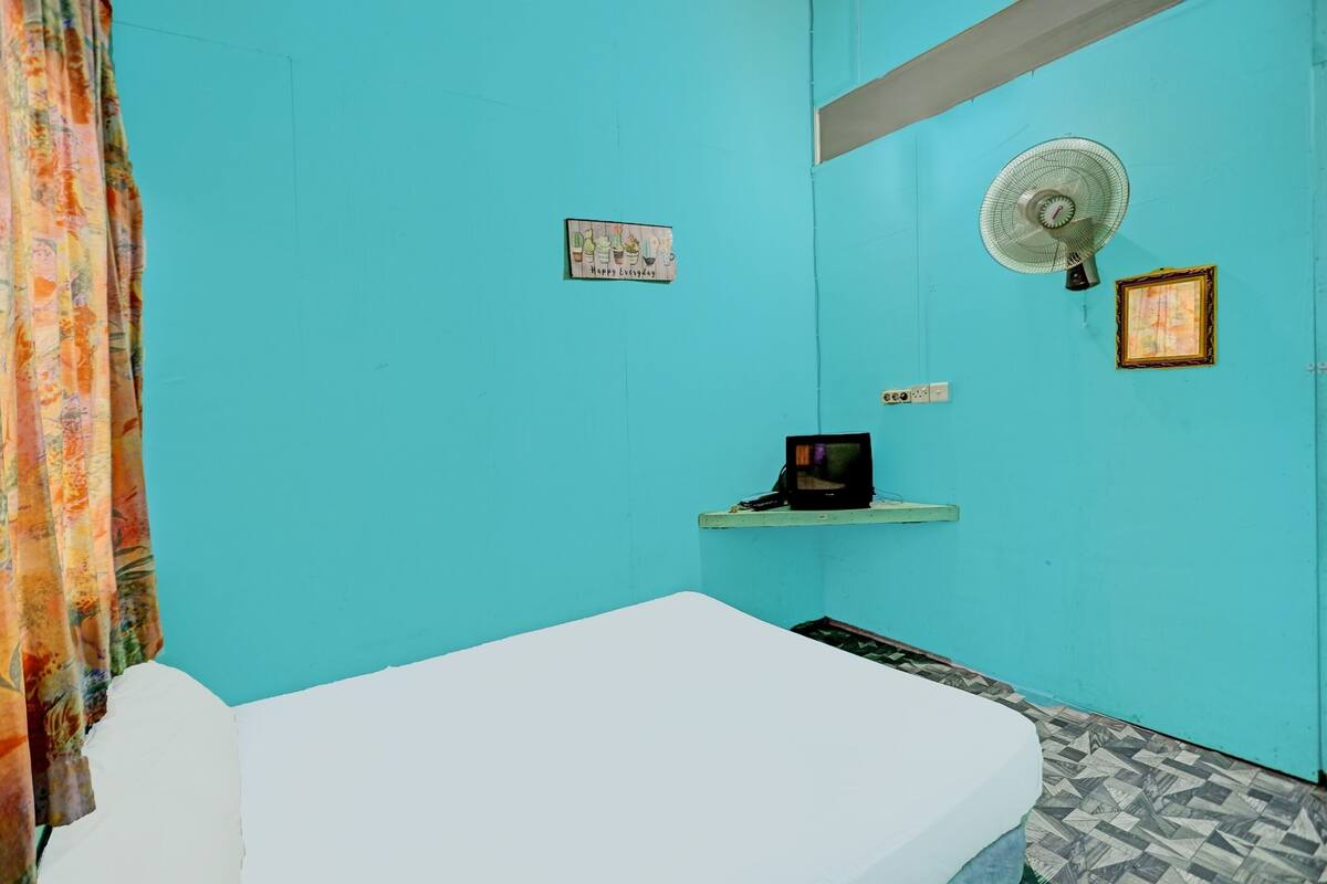 A room with a white bed, a teal wall, a fan, a small shelf with a picture, and a curtain.