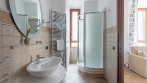 A modern bathroom with a large oval mirror, a double sink, a glass-enclosed shower, and a beige sofa in the adjacent living area. - Flaminiarooms (Rome)