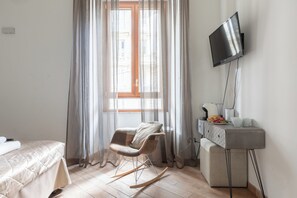 Une chambre d’hôtel comprenant un lit, un fauteuil à bascule, un bureau avec un téléviseur à écran plat et une fenêtre avec des rideaux légers.