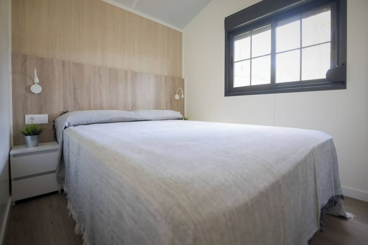 Un dormitorio con una cama, una mesita de noche con una planta, una lámpara de pared y una ventana con persianas.