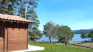 A wooden cabin with a tiled roof overlooking a lake, surrounded by trees and a clear sky. - Camping Urbión (Soria)