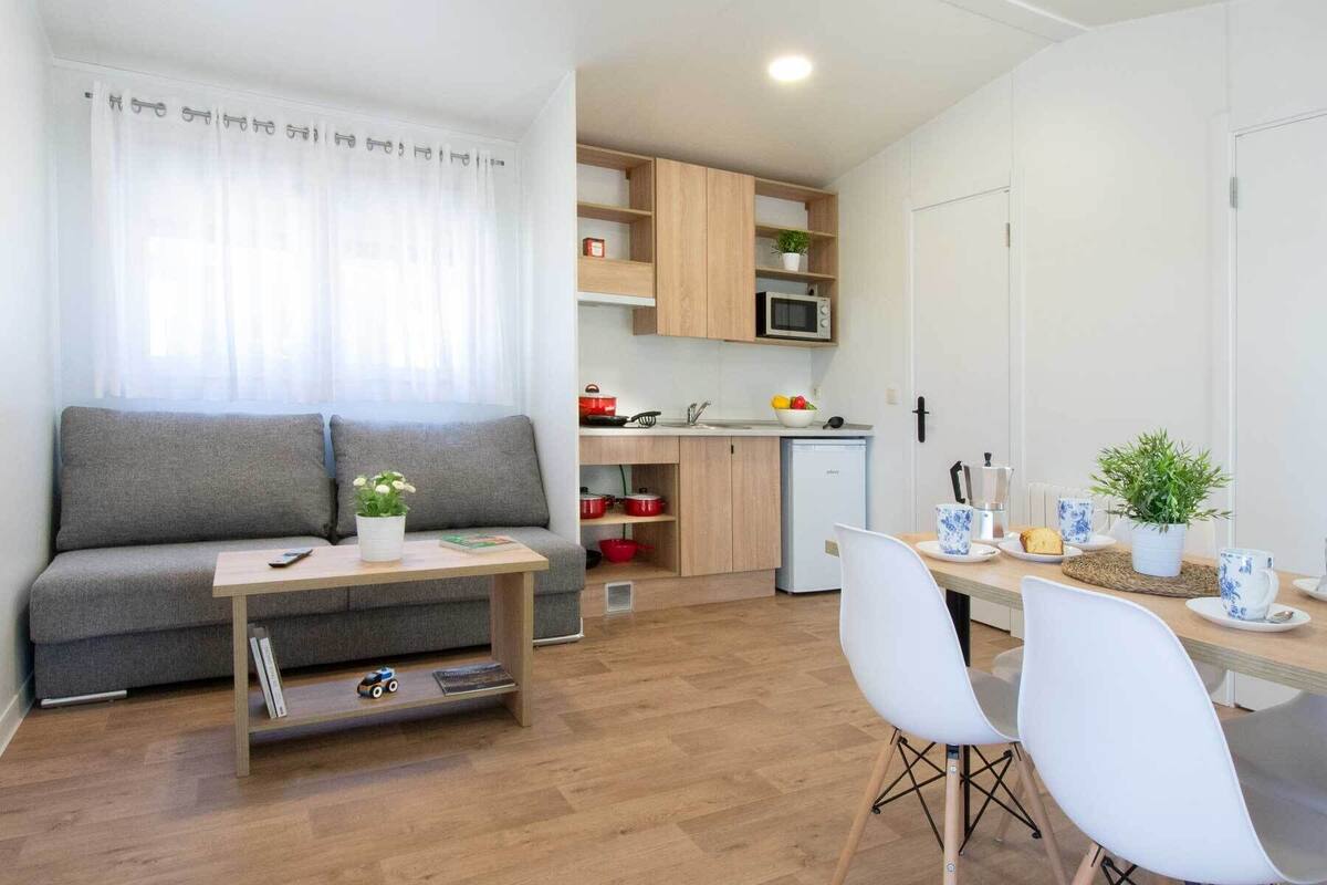 Un espacio habitable compacto con un sofá gris, una pequeña mesa de comedor con sillas blancas, una kitchenette con microondas y una planta en maceta.