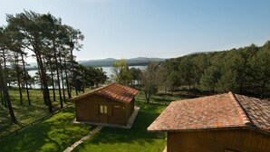 A wooden cabin with a tiled roof, surrounded by a well-maintained lawn and trees, with a lake and mountains in the background. - Camping Urbión (Soria)