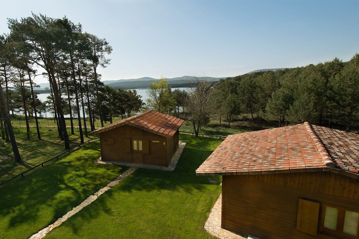 Una cabaña de madera con techo de tejas, rodeada de un césped cuidado y árboles, con un lago y montañas al fondo.