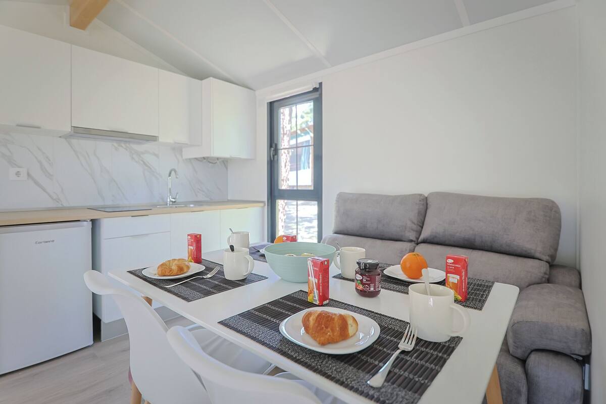 Una cocina moderna con armarios blancos y un protector contra salpicaduras de mármol. Hay una mesa de comedor con dos platos de croissants, una taza de café y un zumo. Un sofá gris se ve al fondo.