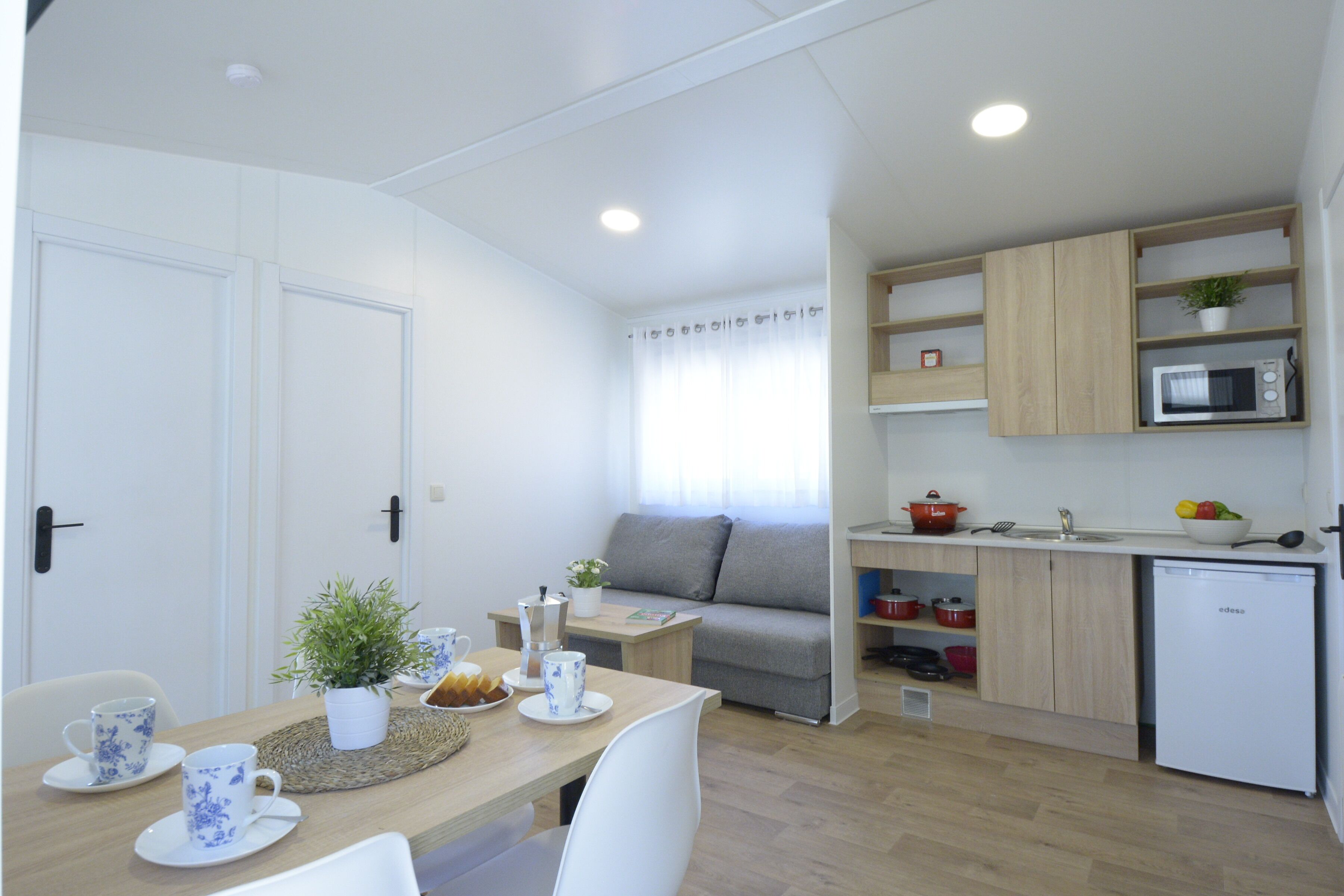A compact kitchen with a microwave, sink, and wooden cabinets. There is a dining table with chairs and a small plant on the table.