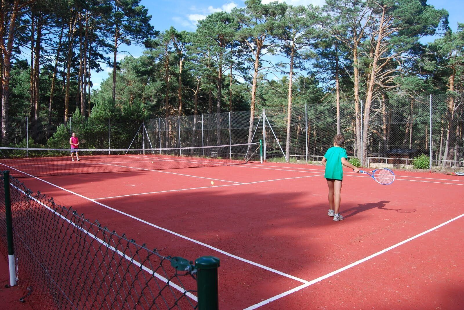 A tennis court with two players, surrounded by a chain-link fence and tall trees.