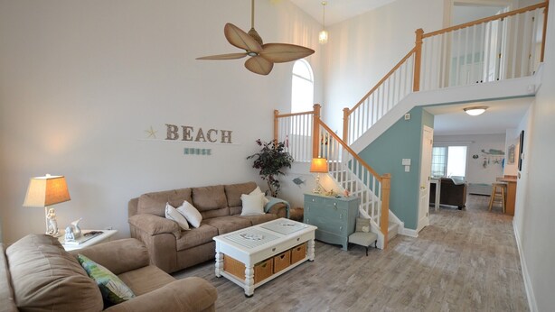 A spacious living room with a brown sofa, a white coffee table, and a ceiling fan. A staircase leads to an upper level.