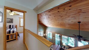 Un dormitorio estilo loft con techo alto, vigas de madera, cama con ropa de cama a rayas, ventilador de techo y vistas a un balcón con paisaje exterior.
