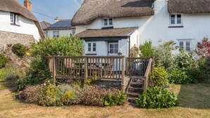 Une maison de campagne traditionnelle avec un toit de chaume, une terrasse en bois et un jardin où poussent différentes espèces de plantes.