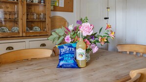 Une table de salle à manger en bois, ornée d’un vase de fleurs, d’une bouteille de jus de fruits et d’un paquet de chips légèrement salées.