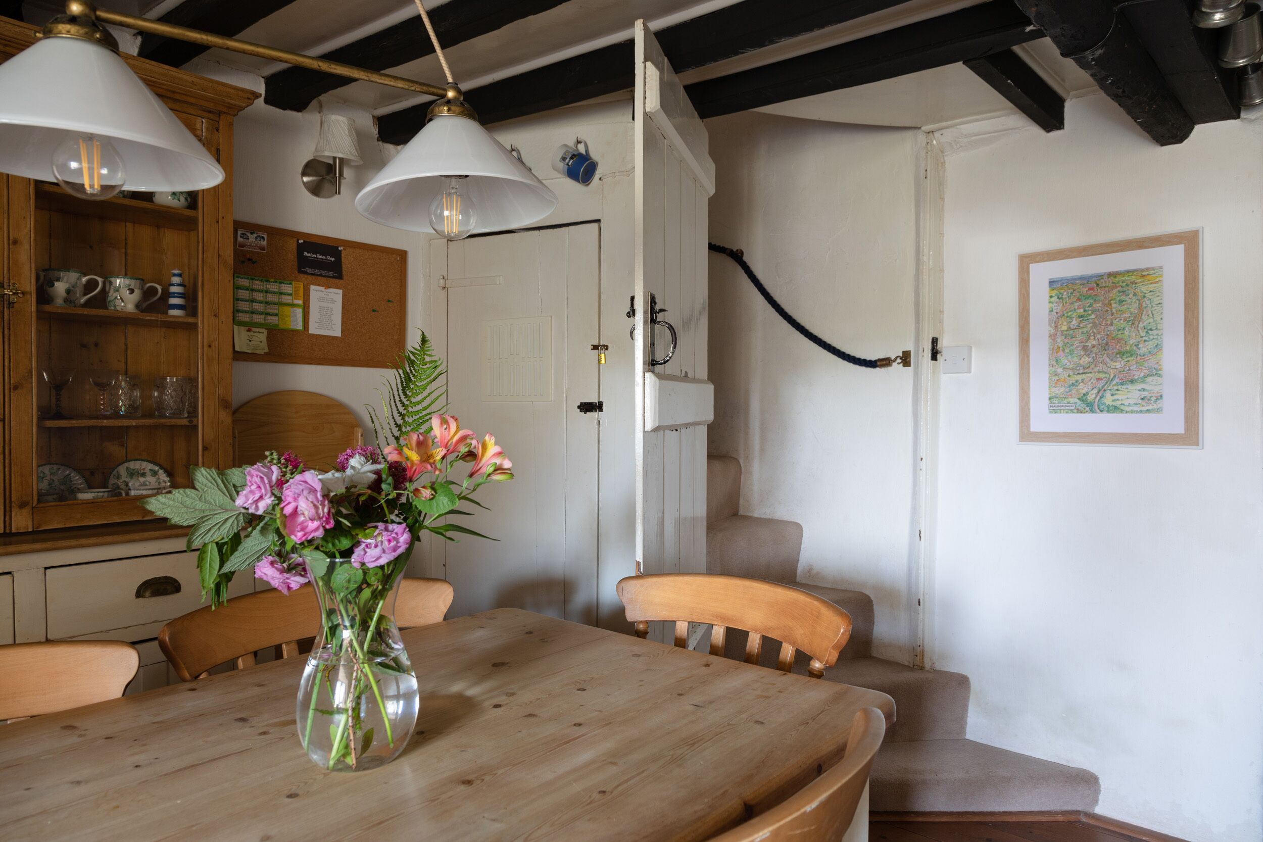 Una mesa de comedor de madera con sillas, un jarrón de flores y un tablón de anuncios con notas y un mapa en la pared.