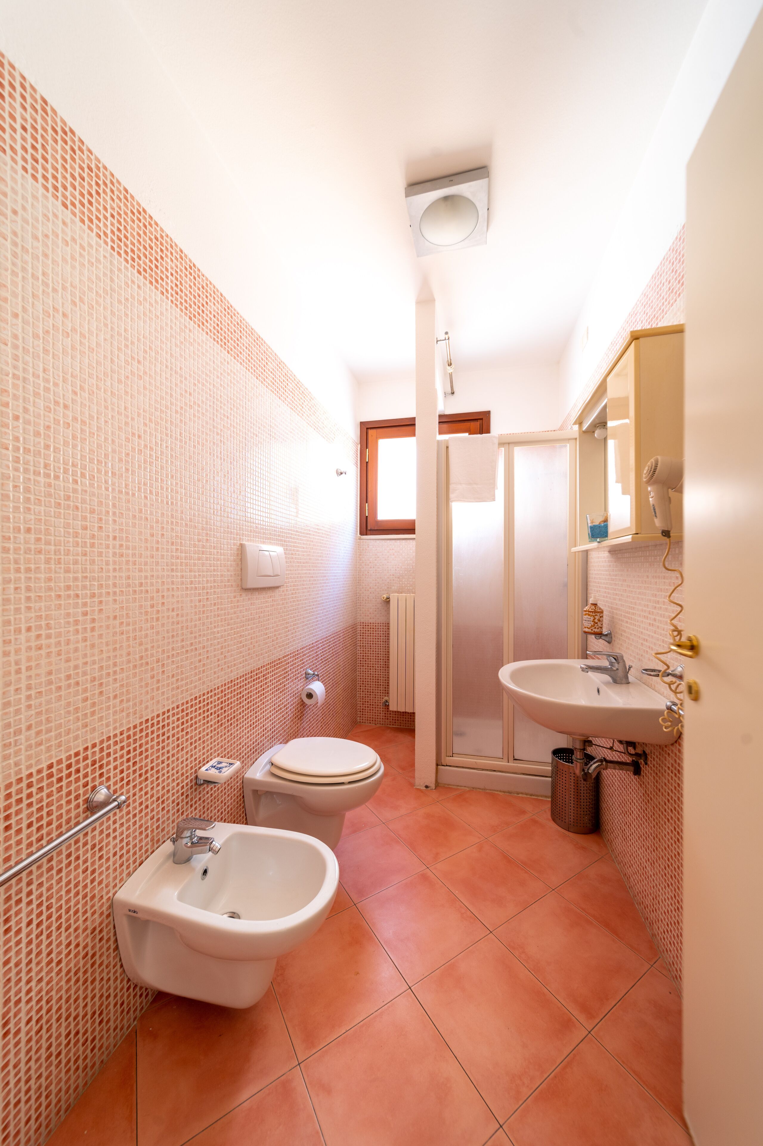 Un baño con bidé, inodoro y lavabo, con azulejos en una cálida gama de colores, una puerta de ducha y un perchero para toallas.