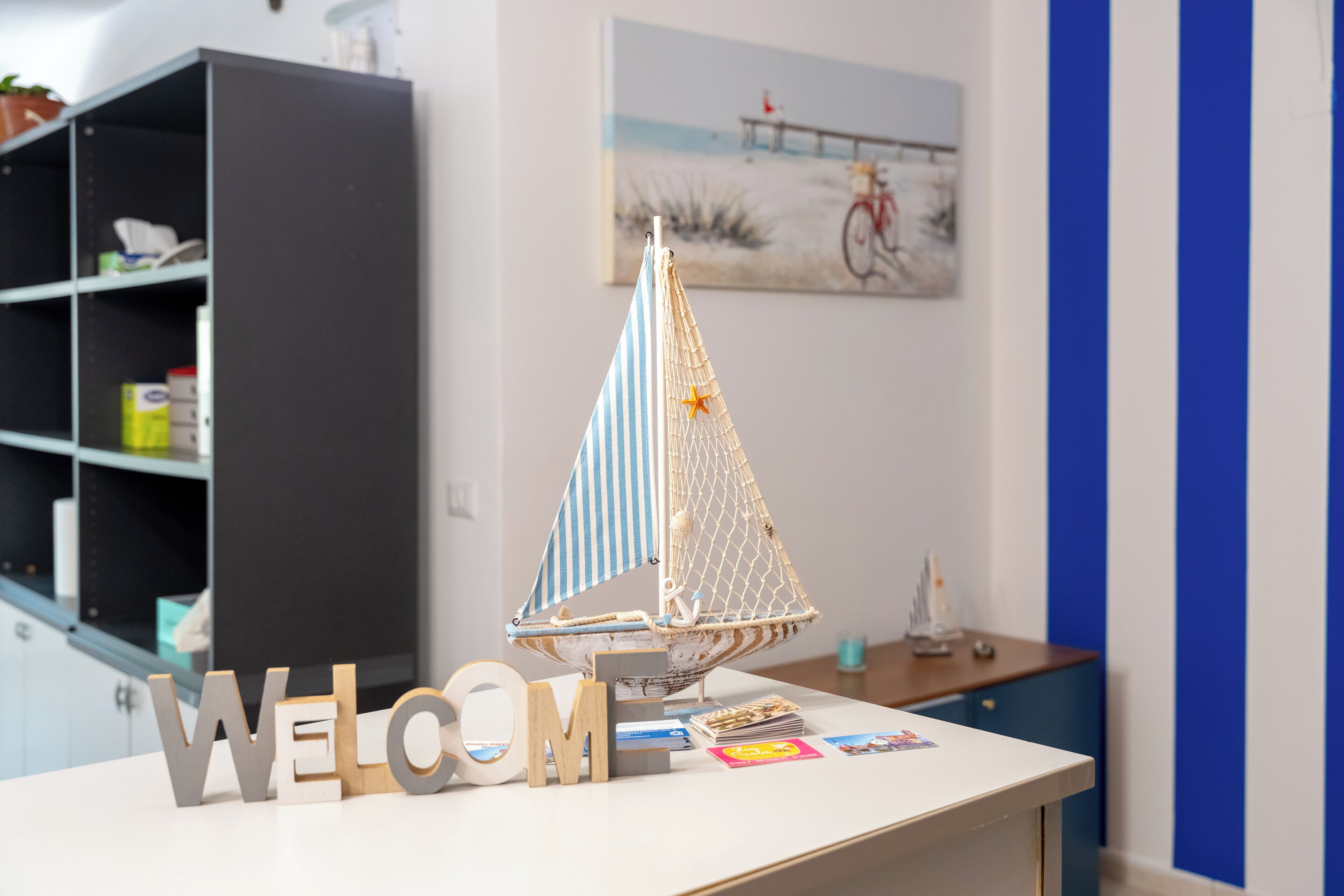 A model sailboat on a desk with 'WELCOME' letters, a bookshelf, and a painting in the background.