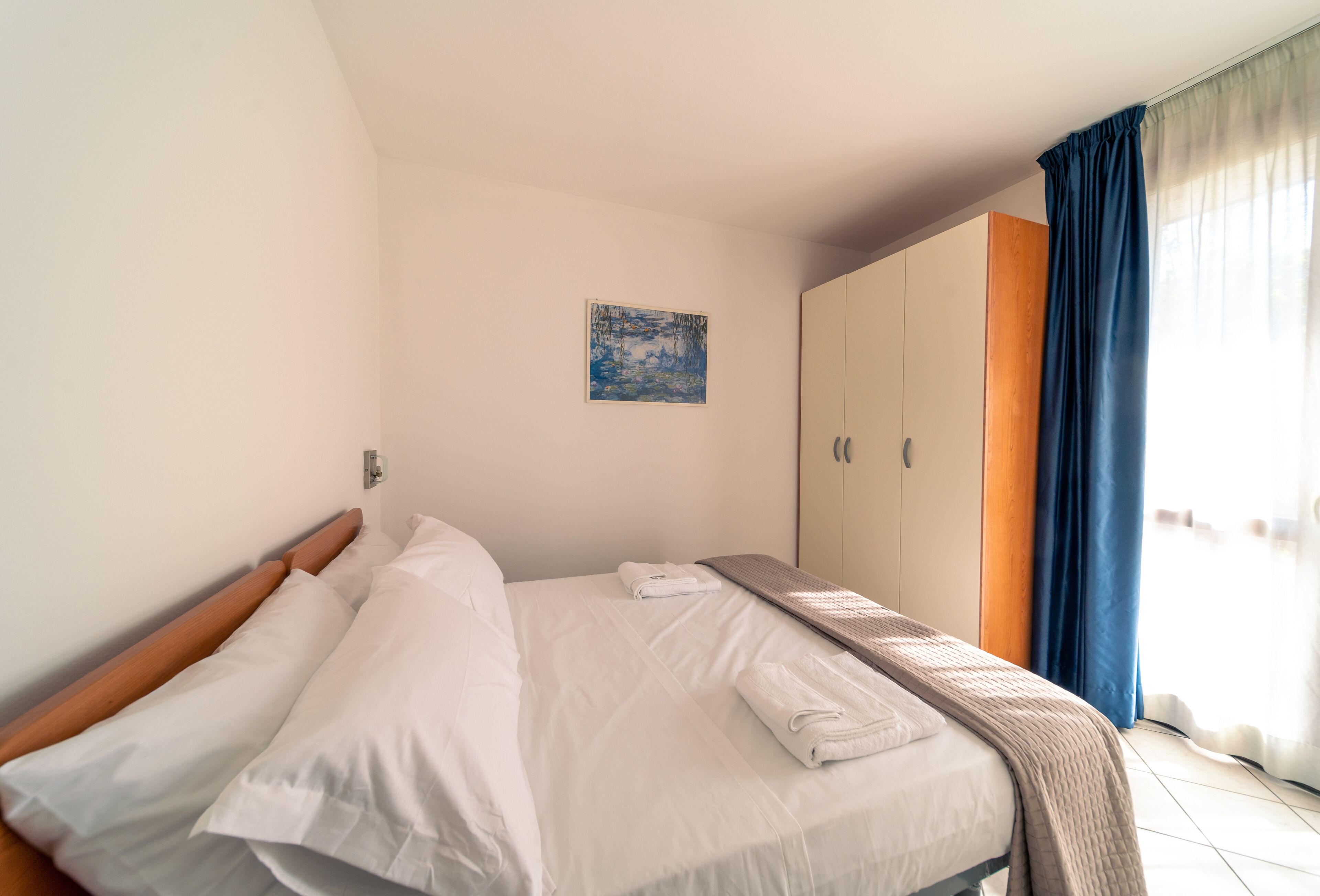 A neatly made bed with white linens, a blue artwork on the wall, and a wardrobe with a wooden panel.