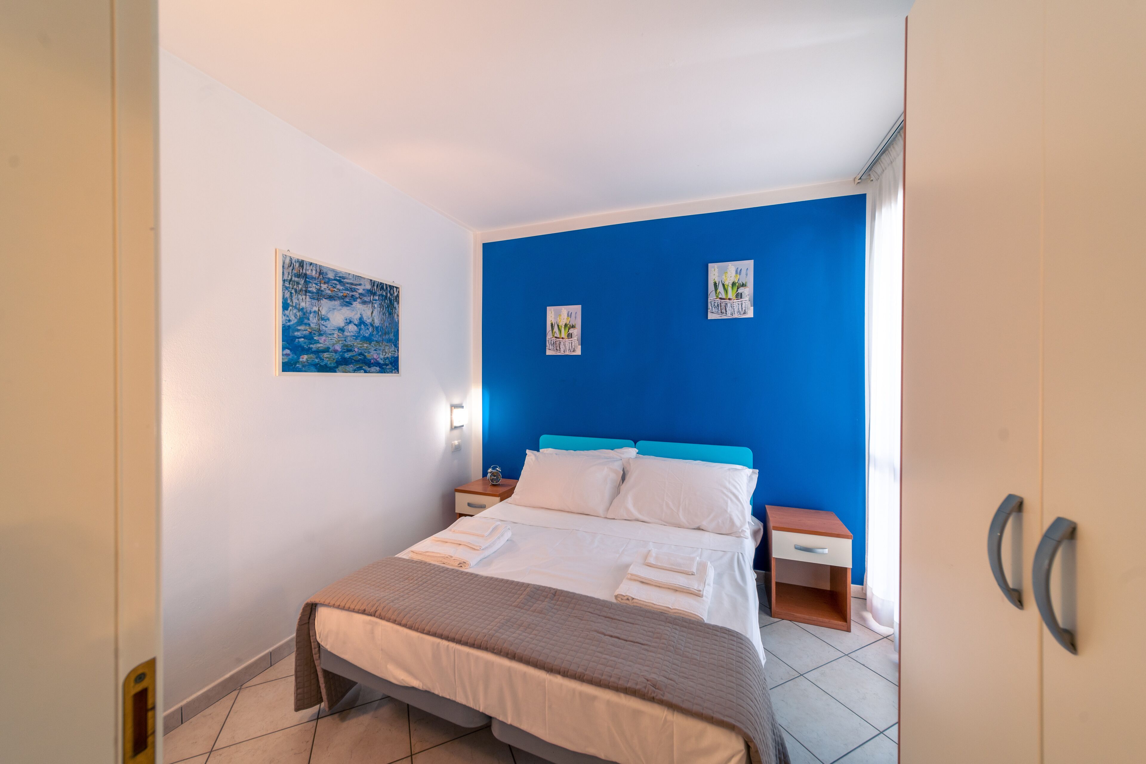 Una cama bien hecha con sábanas blancas, un cabecero azul y una mesita de noche con una lámpara.