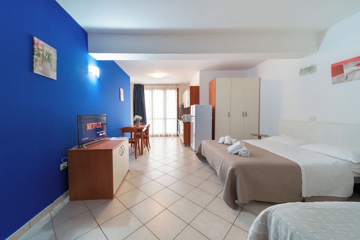 A hotel room with a bed, a desk with a computer, a TV displaying Netflix, a window with curtains, and a kitchenette area.