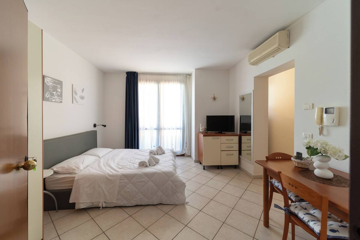 A hotel room with a bed, a desk with a chair, a TV, a phone, and a window with curtains.