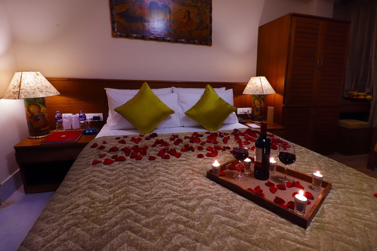 A hotel room with a bed, two green pillows, a bedside table with a lamp, a laptop, and a tray with rose petals, candles, and wine glasses.