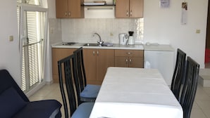 Eine kleine Küche mit Holzausstattung, einer Spüle, einer Kaffeemaschine und einem Kühlschrank. Es gibt einen Esstisch mit Stühlen und ein Fenster mit Jalousien.