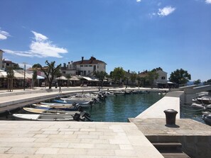 A marina with boats docked, a paved walkway, and buildings with outdoor seating. - Apartment for 3–4 people with a magnificent view of the sea (Njivice)