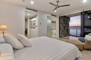 Une chambre avec un lit, un ventilateur de plafond, une vue sur la mer par une porte vitrée et un balcon doté de meubles en osier.