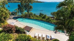 A swimming pool with clear blue water, surrounded by lounge chairs and greenery, overlooking a calm sea and distant hills. - Edge Apartment 15, Edge Apartment 15 on Hamilton Island (Hamilton Island)