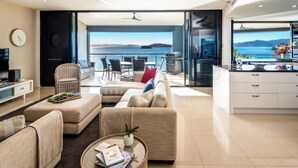 A modern living room with a sofa, armchairs, a coffee table, and a dining area with chairs. A television is on a stand, and there is a view of the sea through large windows. - Edge Apartment 15, Edge Apartment 15 on Hamilton Island (Hamilton Island)