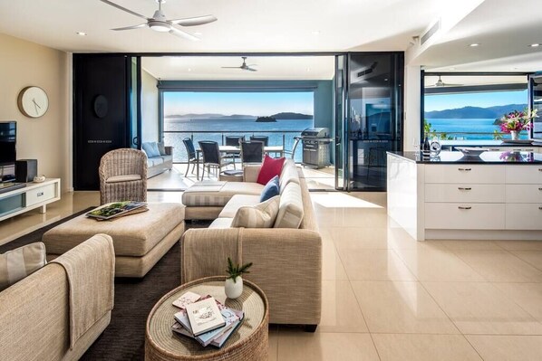 Un salon moderne avec un canapé, des fauteuils, une table basse et un coin repas avec des chaises. Un téléviseur est posé sur un meuble, et de grandes fenêtres offrent une vue sur la mer.
