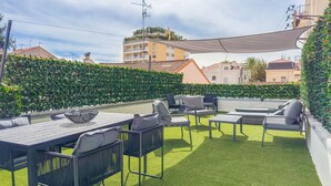 An outdoor patio with a dining table, chairs, and a sofa. There is a hedge and a building in the background. - Le Riviera (Cannes)