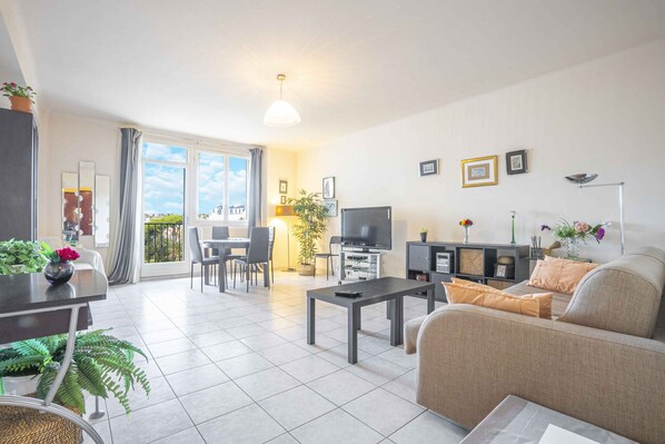 A modern living room with a sofa, coffee table, TV, and dining area. There is a balcony with a view. - Le Molière (Cannes)