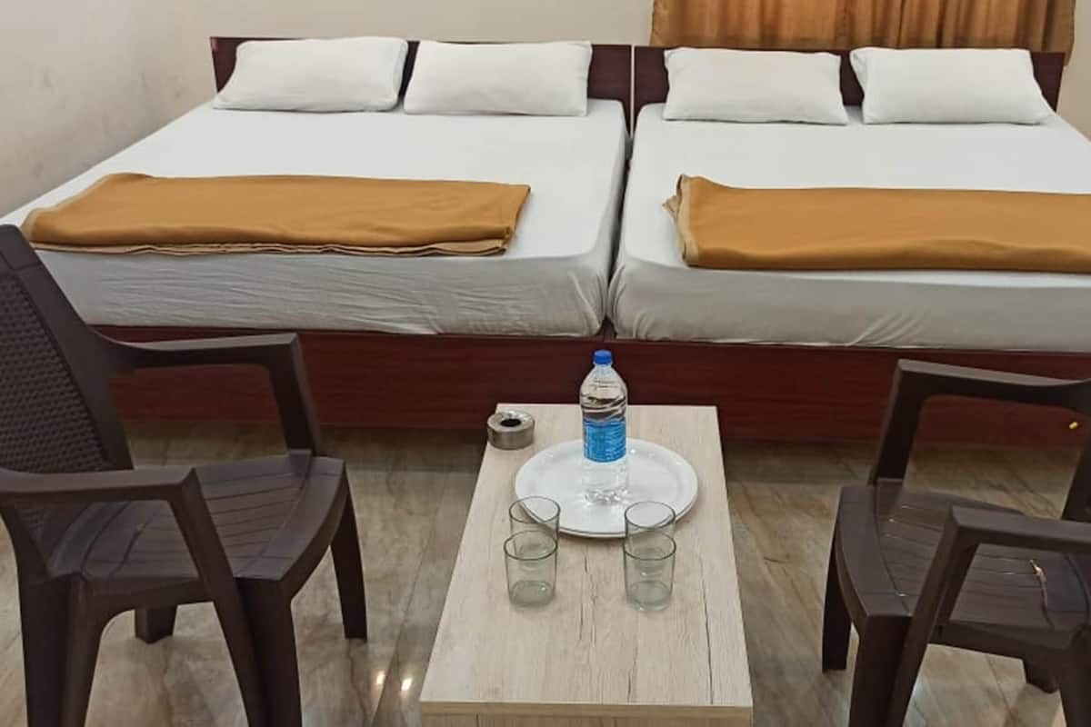Uma cama de casal com duas almofadas e um cobertor marrom, uma pequena mesa com uma garrafa de água e duas cadeiras.